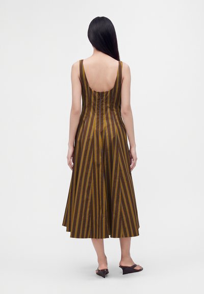 Femme aux longs cheveux noirs portant une robe midi sans manches à rayures verticales marron et jaune, vue de dos, debout sur un fond uni.