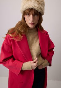 Rózsaszín gyapjú kabát széles gallérral egy bézs kötött pulóver fölött. A modellen egy bolyhos krémszínű kalap van, világos semleges háttér előtt.