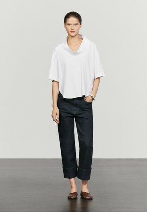 Frau steht und trägt ein weites weißes Kurzarmshirt, dunkle hochgekrempelte Jeans, braune flache Schuhe und ein silbernes Armband am linken Handgelenk.