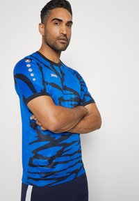 Blaues Sporthemd mit schwarzem abstraktem Muster, kurzenÄrmeln und rundem Halsausschnitt. Mit Logo und dekorativen Knöpfen an der Schulter.