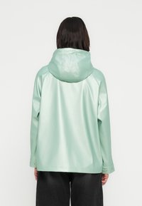 Μέντα πράσινο, αδιάβροχο μπουφάν με κουκούλα, γυαλιστερή υφή, μανίκια ράγκλαν και λεπτομέρειες με κουμπιά στο μανίκι. Εμφανίζεται η οπίσθια όψη.