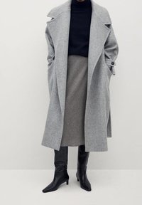 Mujer vestida con un abrigo largo gris, un suéter negro de cuello alto, una falda de cuadros y botas de tacón negras, de pie frente a un fondo sencillo.