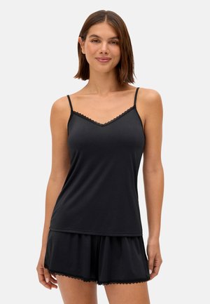 Mujer con camisola negra con encaje y pantalones cortos a juego, de pie contra un fondo claro y liso, sonriendo suavemente.