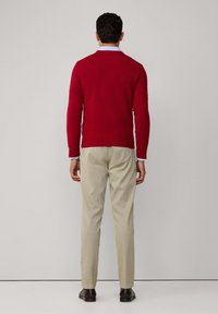 Homme portant un pull rouge, une chemise bleu clair, un pantalon beige et des chaussures marron foncé, se tenant face à un mur gris clair uni.