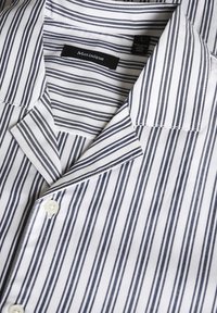 Camisa blanca de botones con rayas verticales en azul marino, y un cuello con muesca, además de un bolsillo en el pecho. La etiqueta dice "Martinique". El tejido parece suave y ligero.