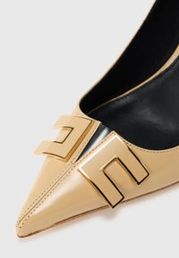 Μπεζ παπούτσι με ψηλό τακούνι από δέρμα λουστρίνι, με μυτερή μύτη, που διαθέτει έντονη χρυσή γεωμετρική αγκράφα και μαύρο εσωτερικό.