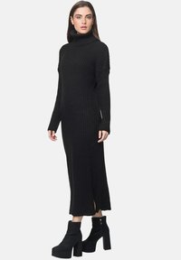 Schwarzes, geripptes Strick-Turtleneck-Kleid mit langen Ärmeln und seitlichen Schlitzen. Kombiniert mit schwarzen, hochhackigen Stiefeletten.