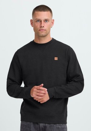 Jonge man met kort haar die een zwart sweatshirt met lange mouwen draagt, met een klein bruin vierkant logo op de borst, staande met de handen in elkaar.