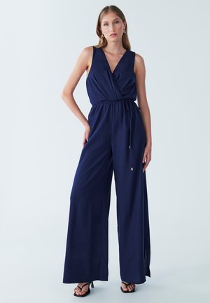 Combinaison bleu marine avec un design croisé, jambes larges et taille à cordon. Présente un décolleté en V profond et des fentes latérales. Tissu lisse et léger.