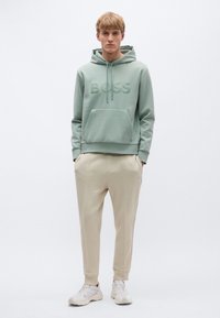 Φούτερ με κουκούλα σε μέντα πράσινο χρώμα με ανάγλυφο λογότυπο "BOSS", τσέπη καγκουρό, συνδυασμένο με μπεζ κωνικά φούτερ παντελόνια και λευκά αθλητικά παπούτσια. Ομαλή υφή.