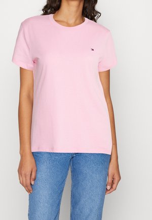 Kobieta nosząca jasnoróżowy t-shirt z krótkim rękawem z małym wyszywanym logo na piersi oraz niebieskie jeansy z denimu.