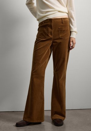 Pantalones de pana marrones de corte acampanado con bolsillos traseros, combinados con un suéter de color claro y zapatos mocasines oscuros, sobre un fondo neutro.