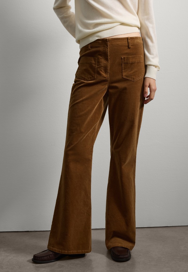 Braune Corduroy-Hosen mit ausgestelltem Bein und hinteren Taschen, kombiniert mit einem hellen Pullover und dunklen Mokassinschuhen, vor einem neutralen Hintergrund.
