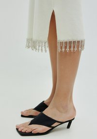 Witte jurk met een split, gedetailleerd met zilveren kralenfranje aan de onderrand. Zwarte teenslippers met een triangulaire design en een lage hak.