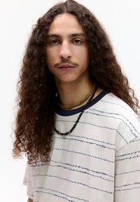 Hvid t-shirt med blå vandrette striber, rund marineblå halsudskæring. Modellen har langt krøllet hår og bærer lagdelte perlehalssmykker.