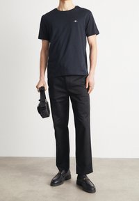 T-shirt nera girocollo con un piccolo logo sul petto, abbinata a pantaloni neri larghi e mocassini neri, con una borsa in mano.