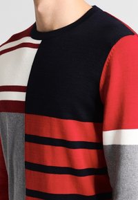 Pull en maille avec un col rond, sections multicolores en bleu marine, rouge, gris et blanc, avec des rayures horizontales et une texture douce.