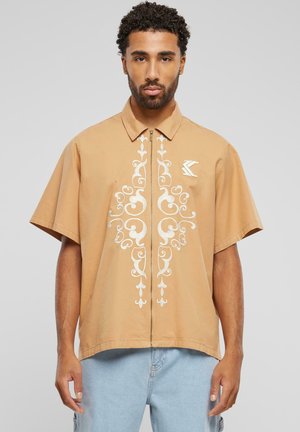 Chemise zippée beige à manches courtes, présentant des motifs floraux brodés blancs au centre et un petit logo sur la poitrine.