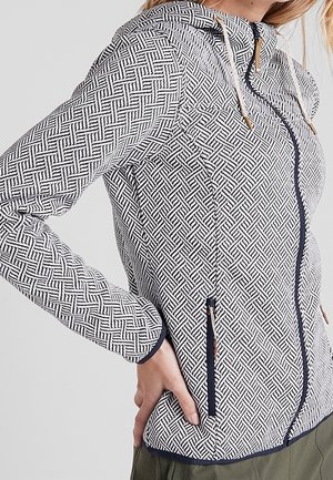 Kvinde iført en hvid og sort mønstret hættetrøje med lynlås, forlomme og snore foran mod en ensfarvet baggrund.