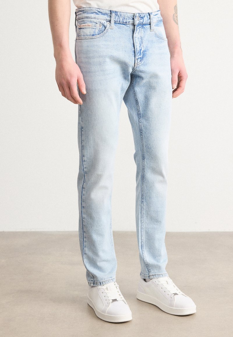 Mężczyzna w jasnoniebieskich obcisłych dżinsach i białych trampkach, stojący na szarej podłodze na tle gładkiej białej ściany.