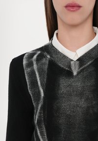 Pulover negru cu un design texturat și dâre albe asemănătoare cu pensula. Decolteu în V cu o cămașă albă în contrast purtată dedesubt.