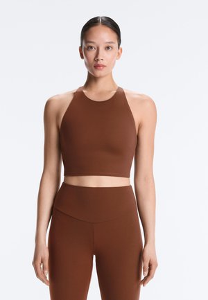 Jonge vrouw met steil donker haar in een strak knotje, gekleed in een bruin mouwloos crop top en high-waisted legging tegen een witte achtergrond.