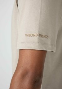 Beige tričko s krátkým rukávem s výšivkou "WRONG FRIENDS" v odstínu tan na levém rukávu; měkká bavlněná tkanina, přiléhavý střih.