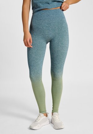 Kobieta ubrana w wysokogatunkowe, bezszwowe legginsy o gradientowym przejściu kolorów od niebieskiego do zielonego, w połączeniu z białymi butami sportowymi, stojąca na jednolitym tle.