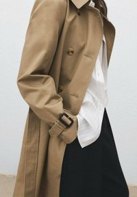 Cappotto trench beige realizzato in un tessuto liscio, con design doppiopetto, bottoni dorati e dettagli sui polsini con cintura.