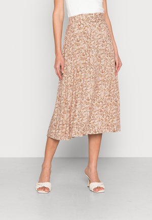 Femme portant une jupe midi fluide taille haute à motif floral rose et blanc, associée à des sandales blanches à talons et tressées.