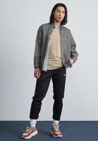 Camisa a rayas verde y gris sobre camiseta beige, pantalones deportivos negros con detalles blancos y zapatillas marrones y crema con suelas texturizadas.