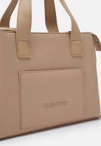 Beige leather handbag with textured surface, featuring dual handles, a front pocket, and embossed brand logo in gold.