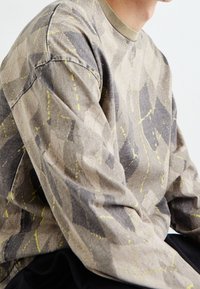 Langarmshirt aus strukturiertem Stoff in Beige mit grauen und leuchtend gelben abstrakten Mustern, mit einem abgerundeten grünen Halsausschnitt.