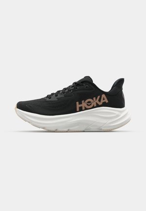 Čierna športová obuv s textúrovaným zvrškom, výrazným logom HOKA a hrubou bielou medzipodrážkou. Má zaoblenú špičku a gumovú podrážku.