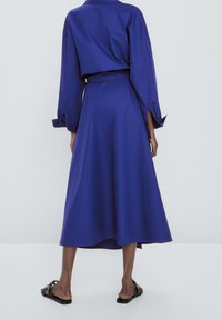 Lång blå klänning med bälte i midjan, vida ärmar och en flödande kjol. Slätt tyg med en lätt lyster, knälång bakhem. Svarta sandaler.