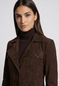 Kvinde med langt mørkt hår iført en mørkebrun ruskindjakke over en sort rullekrave mod en ensfarvet lys baggrund.