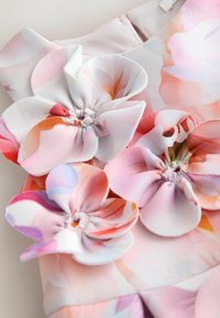 Detalje af kjole i blomstret stof med pink, orange og lilla nuancer, lagdelte stofblomster og en blød, glat tekstur.