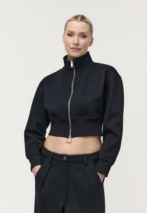 Kvinde med blondt hår iført en sort kort lynlåstrøje og sorte bukser med hænderne i lommerne mod en ensfarvet lys baggrund.