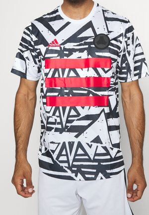 Mężczyzna w czarno-białej koszulce Adidas z geometrycznym wzorem, trzema poziomymi czerwonymi paskami oraz czarnym okrągłym emblematem na piersi.