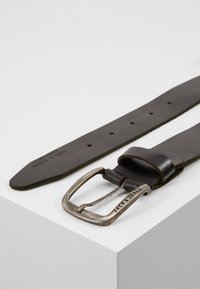 Cinturón de cuero negro con hebilla de metal plateado, que presenta una inscripción de la marca. La correa es plana y tiene agujeros espaciados de manera uniforme para el ajuste.