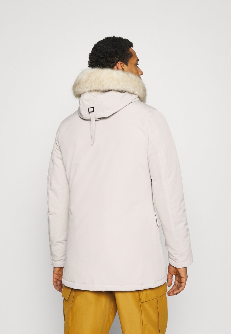 Un homme de dos portant un parka beige clair avec une capuche à fourrure et un pantalon cargo jaune moutarde sur un fond uni.