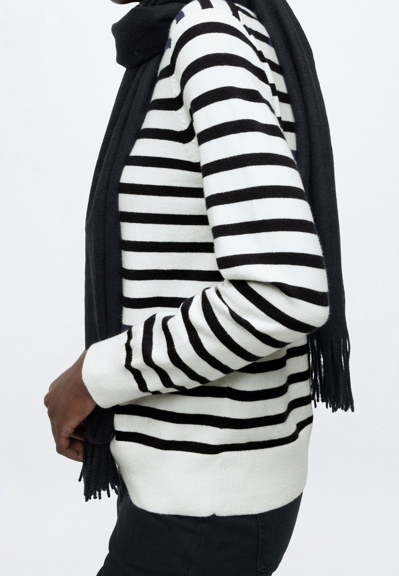 Personne portant un pull à manches longues rayé noir et blanc avec une écharpe noire drapée autour du cou et un pantalon noir.