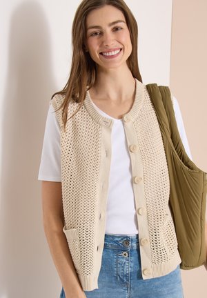 Ung kvinde iført en beige strikket ærmeløs cardigan over en hvid t-shirt, blå jeans og bærer en olivengrøn quiltet jakke.