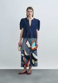 Haut en denim avec manches bouffantes, décolleté en V profond et coutures apparentes ; jupe plissée avec un motif abstrait coloré ; chaussures plates rouges à lacets ; petit sac à main.