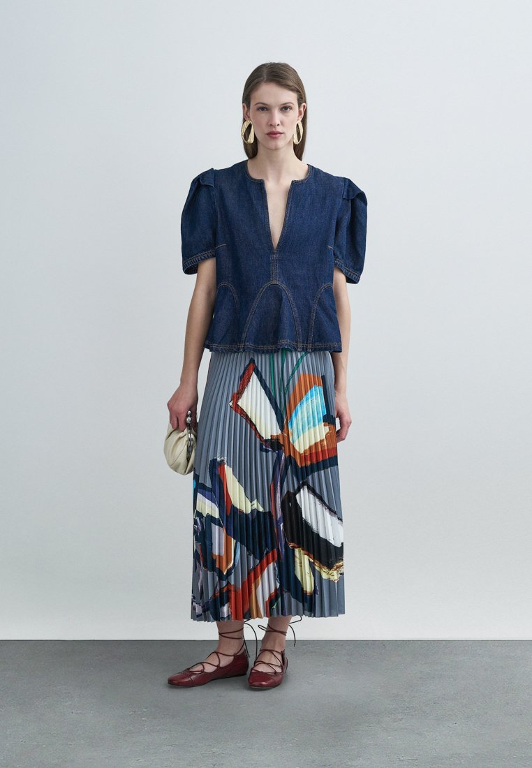 Haut en denim avec manches bouffantes, décolleté en V profond et coutures apparentes ; jupe plissée avec un motif abstrait coloré ; chaussures plates rouges à lacets ; petit sac à main.