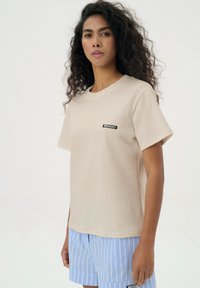 Beżowa bawełniana koszulka z krótkim rękawem, okrągłym dekoltem i czarną naszywką z logo "MISAGA" na lewej piersi. Na dole widoczne jasnoniebieskie spodnie w paski.