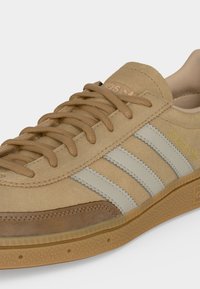 Μπεζ suede αθλητικό παπούτσι Adidas με μπεζ κορδόνια, λευκό σχέδιο με τρεις ρίγες, λαστιχένια σόλα και κείμενο "SPEZIAL" σε χρυσό στη πλευρά.