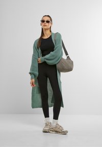 Un cardigan long en tricot ouvert, de couleur vert menthe, porté sur un haut noir ajusté et un legging noir. Accompagné de baskets grises et portant un sac gris matelassé.