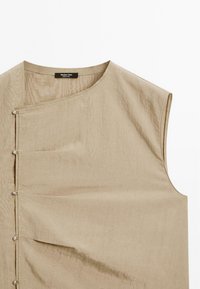 Beige ärmelloses Oberteil mit Rundhalsausschnitt, das über einen diagonalen Verschluss mit metallischen Knöpfen und einem weichen, strukturierten Stoff verfügt.