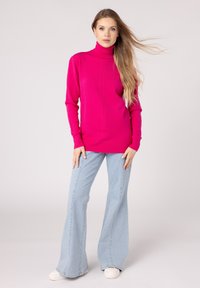 Maglione a collo alto di colore rosa fluo con texture a coste, maniche lunghe e vestibilità rilassata, abbinato a jeans svasati blu chiaro e sneakers bianche.